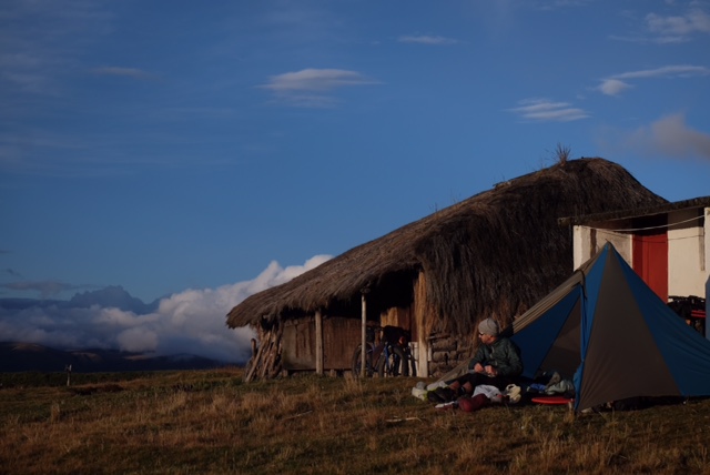A person sitting in front of a tent, on the side of a thatch roof home, in a field, at sunrise