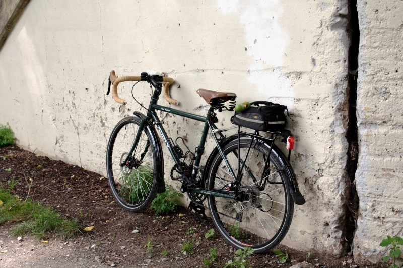 Left side view of a green Surly Disc Trucker bike with a bag on the rear rack, leaning against a white stone wall
