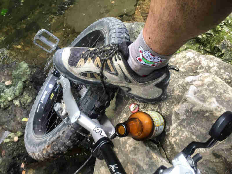 Downward view of a shoed foot, standing halfway on a rock and halfway on a fat bike wheel, next to a beer bottle