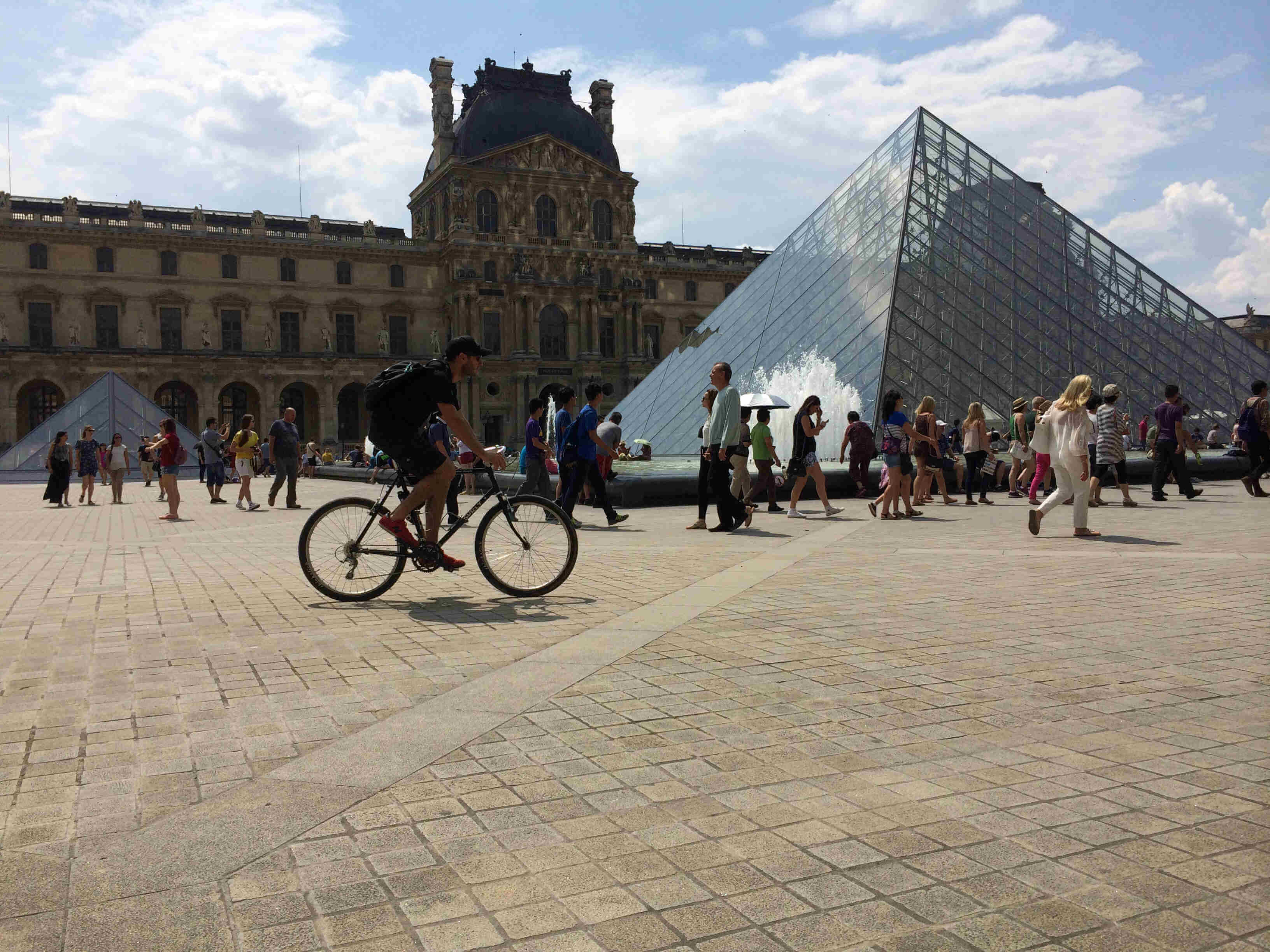 Right side view of a cyclist wearing a backpack, riding a bike across the courtyard in front of the Louvre museum