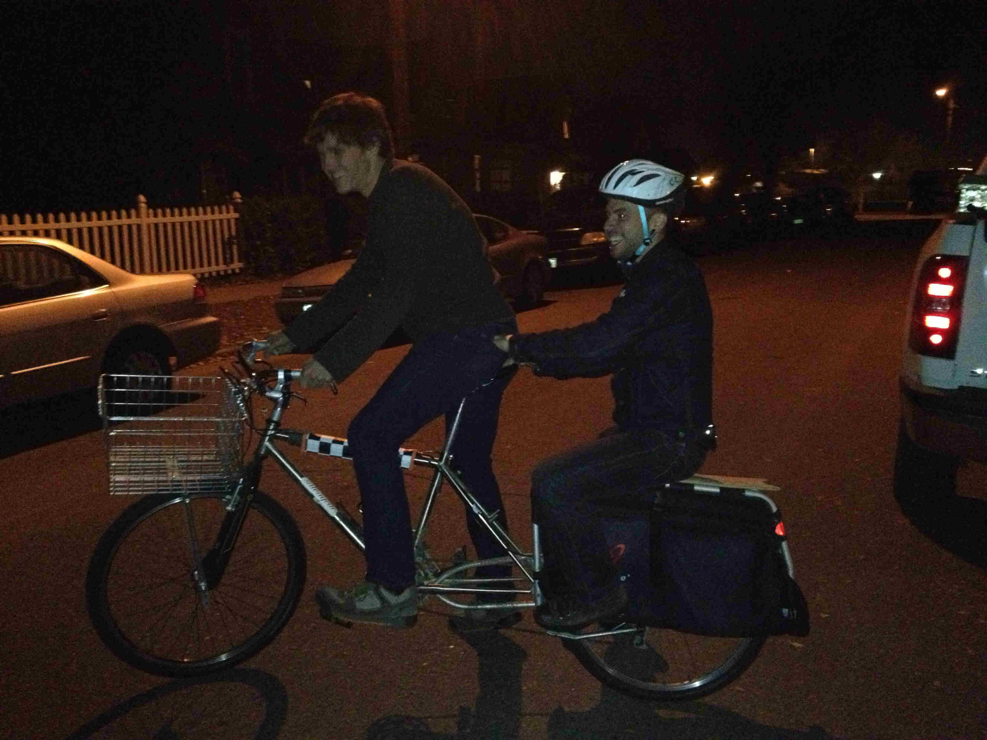Right side view of a silver Surly Big Dummy bike, with a cyclist driving, and a person on back, on a street at night
