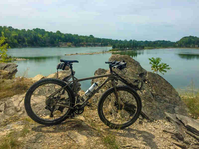 Right side view of a Surly bike, olive, parked on a rock bank, with a lake and trees in the background