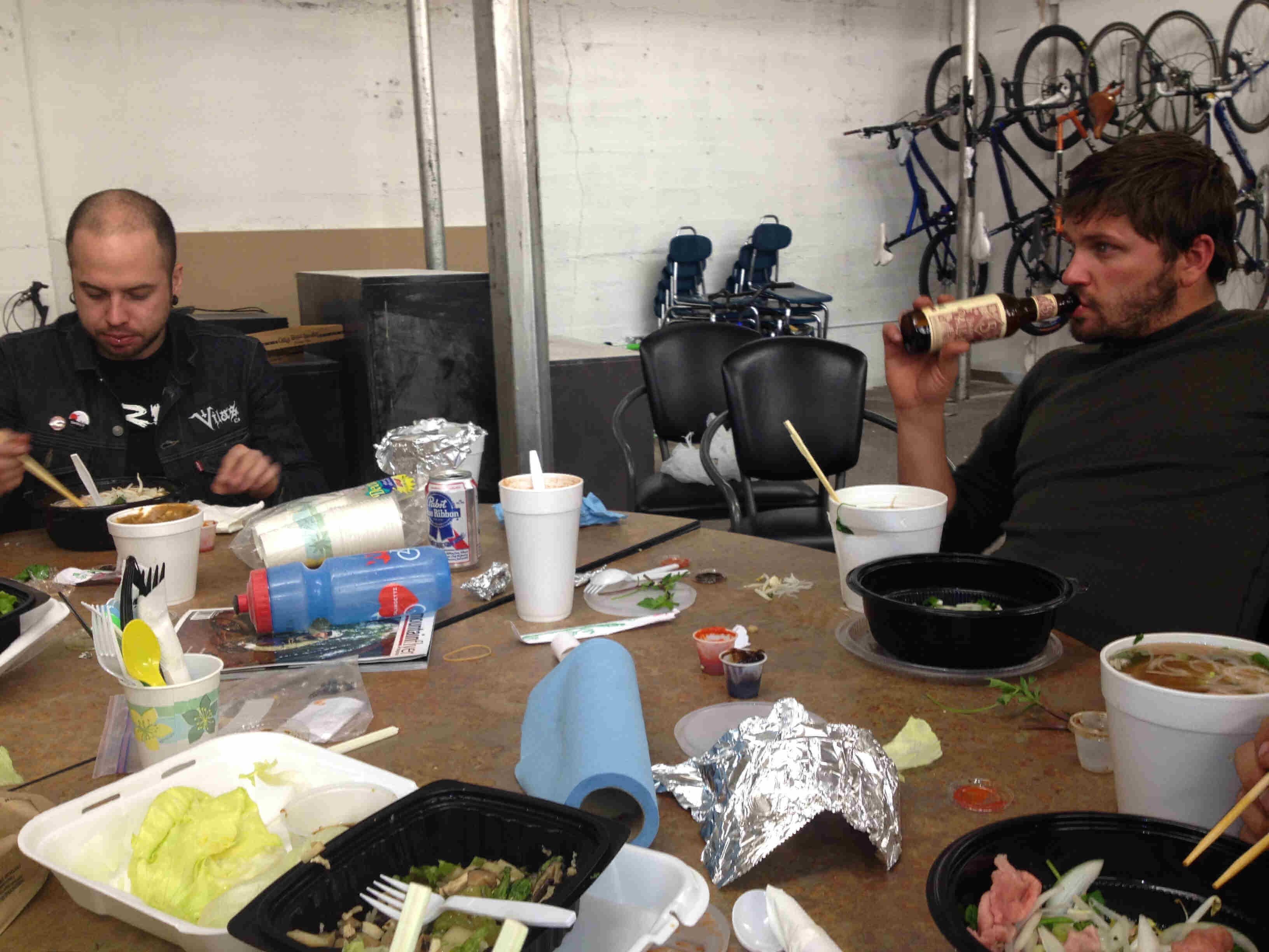 A table full of take-out food, with two people sitting behind it, in a warehouse room with bikes hanging on a wall