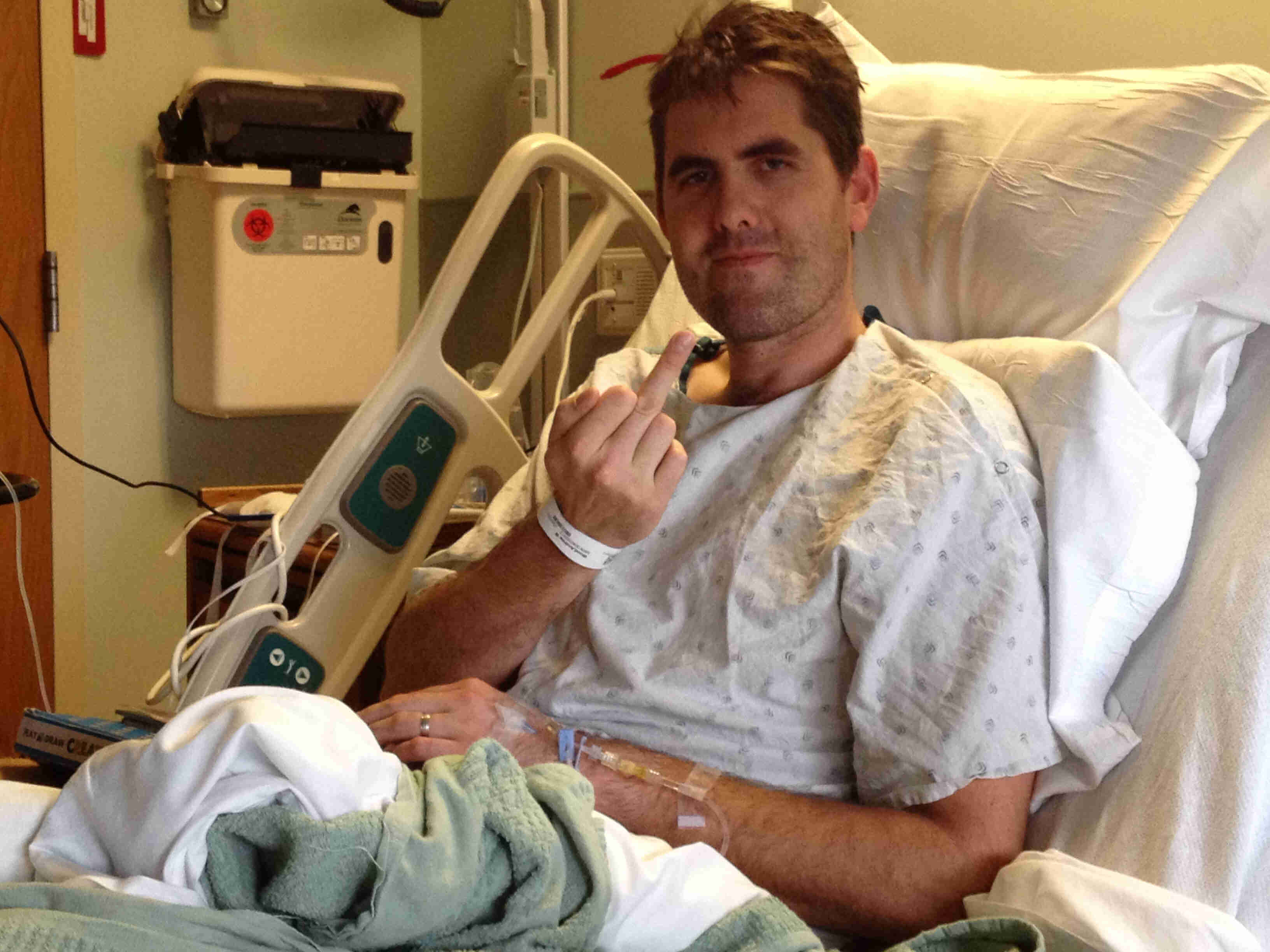 Front left side view of a patient, sitting upright in a hospital bed, holding up their right middle finger