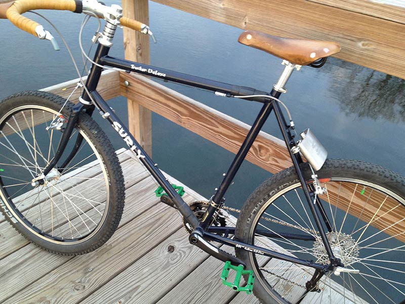 Downward, left side view of a black Surly Trucker Deluxe bike, leaning against a rail on a wood dock, above dark water