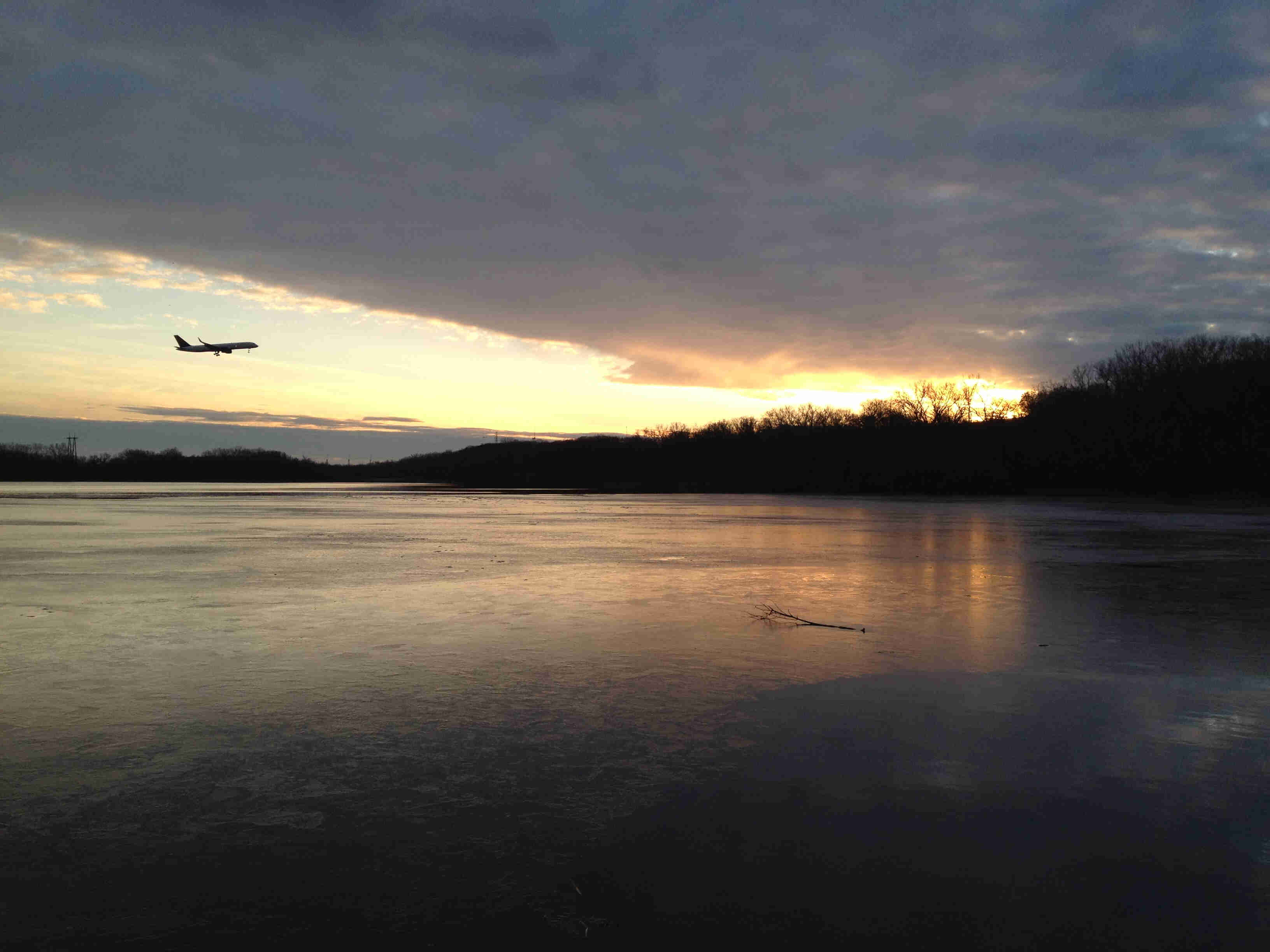 A frozen lake with bare tree around it, with the sun on the horizon and a passenger jet flying across