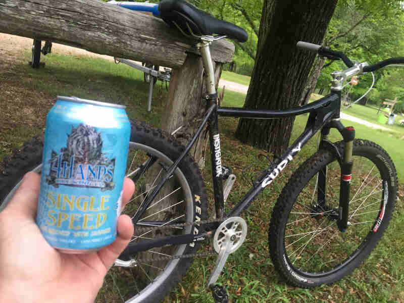 A person's hand holding a beer can in front of a Surly Instigator bike, in a park
