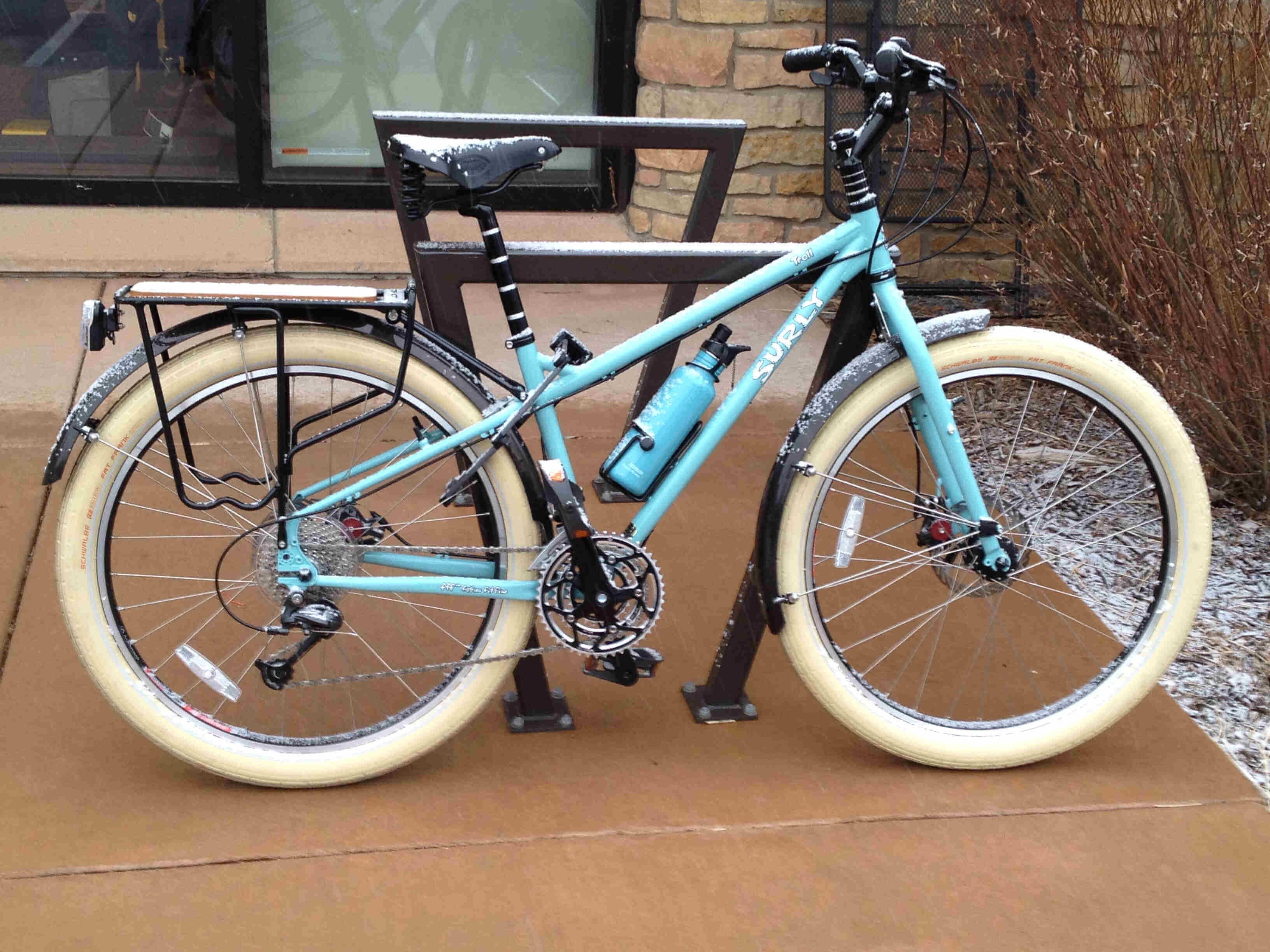 Right side view of a mint Surly Troll bike with white tires, leaning against a bike rack on a sidewalk