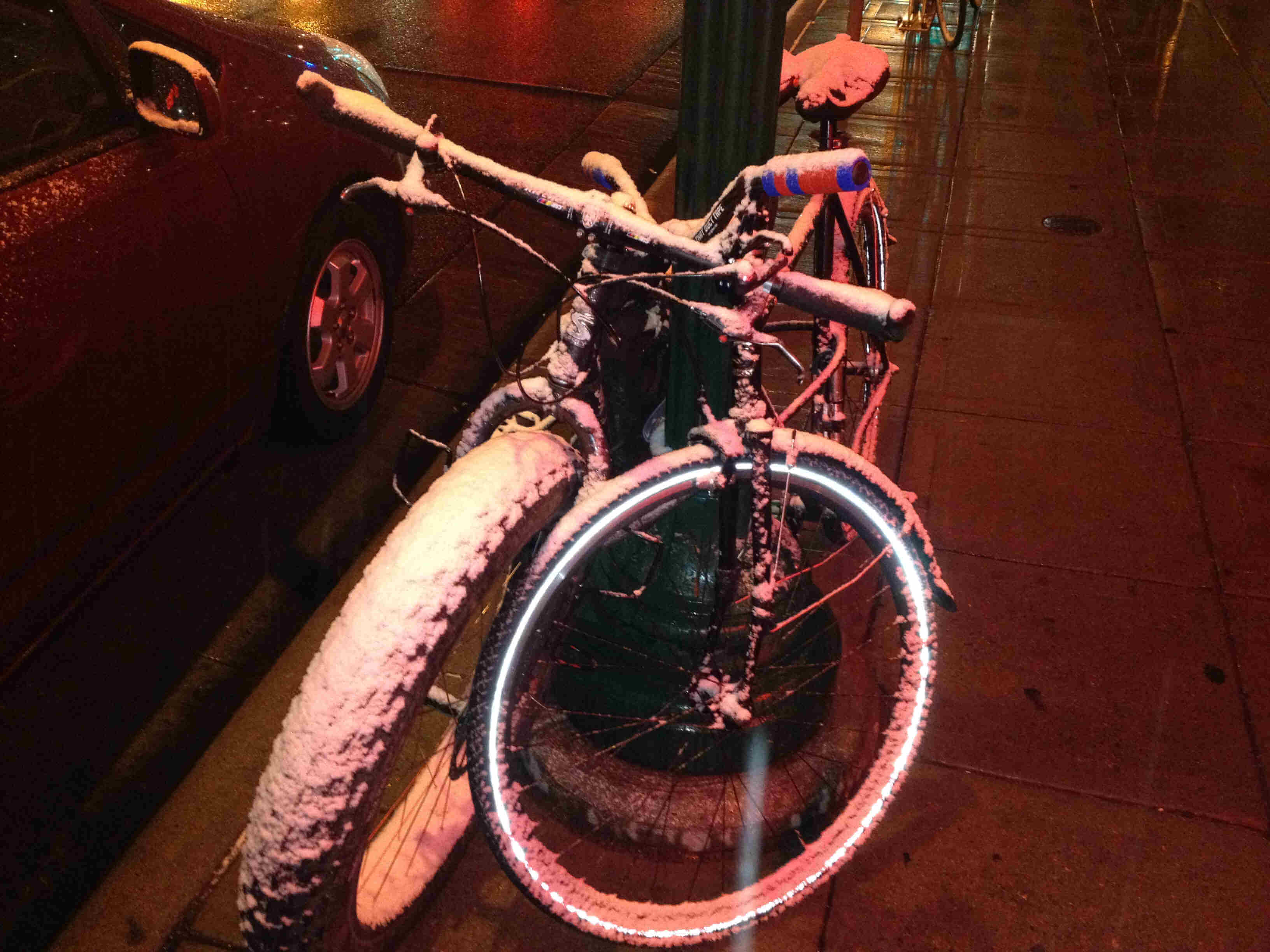 Front view of a fat bike and other bike, coated in snow,  side by side with a light pole between, on a sidewalk at night
