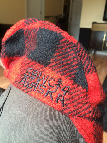 Right side view of a red & black checked cap, with SSWC14 ALASKA embroidered on it, fitted over a person's knee 