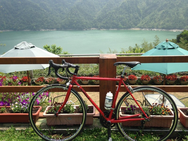 Left side view of a red Surly bike, leaning on a wood fence, with patio behind it, and a mountain lake in the background