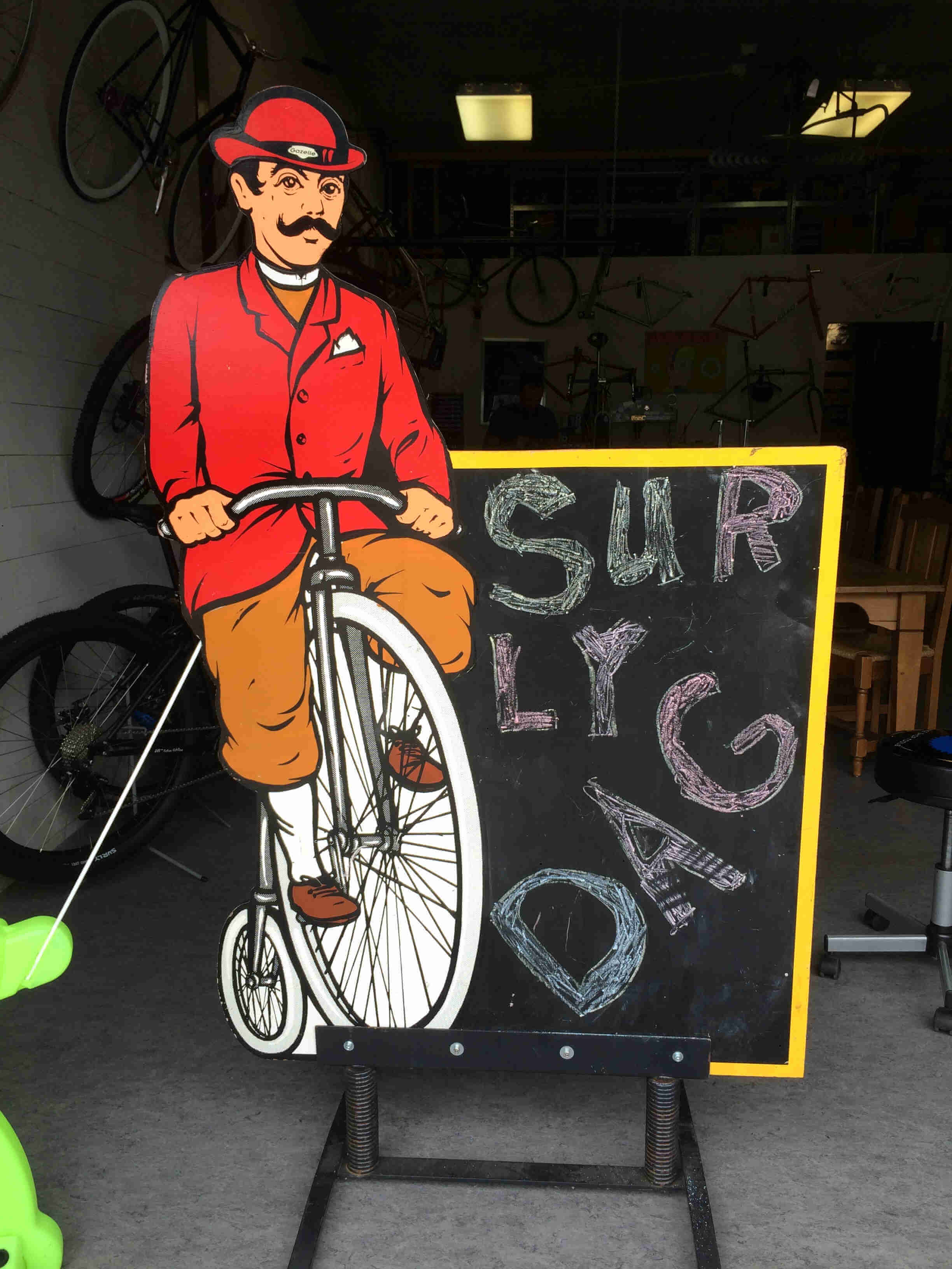 Front view of a sign, with an animated cut out of a cyclist on a high wheel bike, and the words, Surly Dag, written out