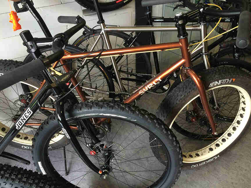 Right side view of copper color Surly Pugsley fat bike, parked between 2 bikes, inside a room with cinder block walls