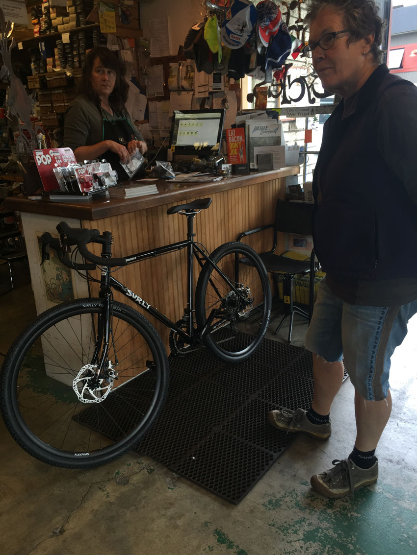 A person standing behind a service counter, helps a customer with a black Surly bike in front of them