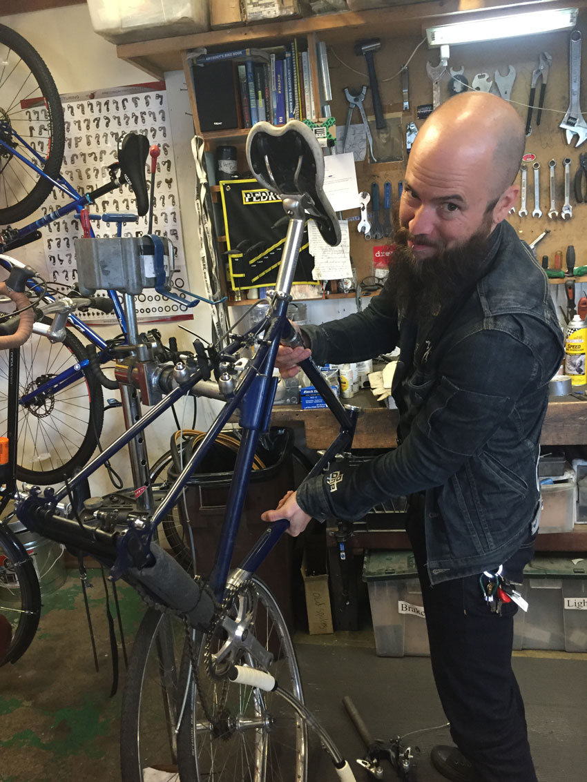 Front view of a person posing, while holding a blue bike on a stand, in a repair shop
