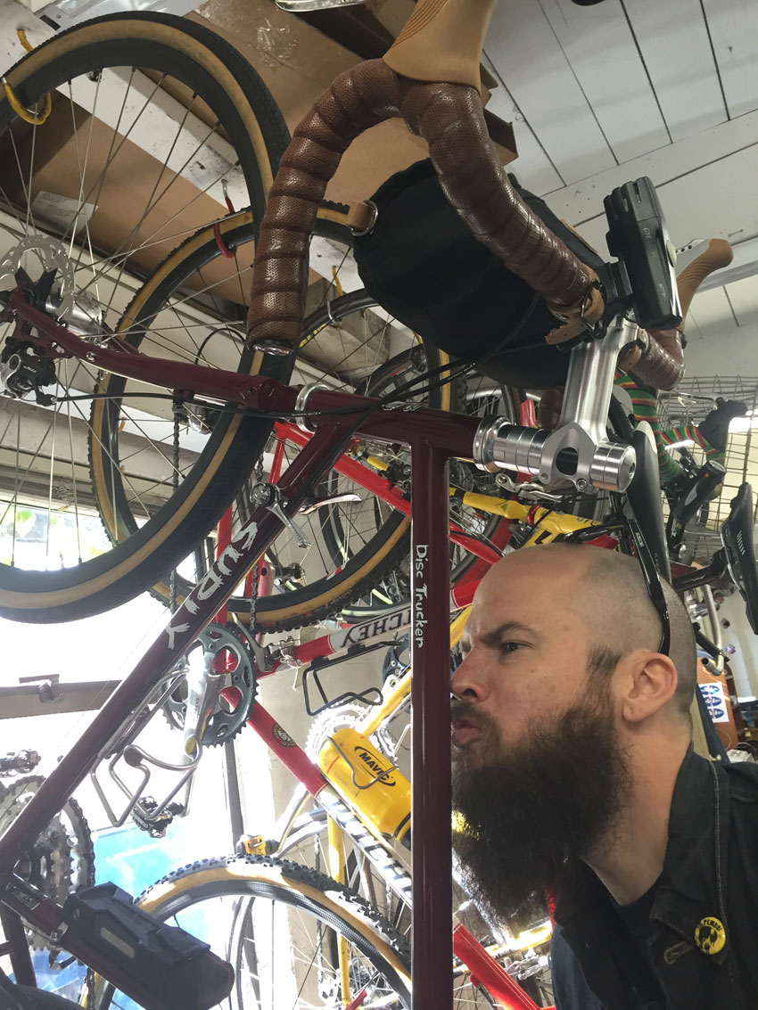 A person with their nose on the top tube of a red Surly Disc Trucker bike, that's hanging from a rack, in a bike shop