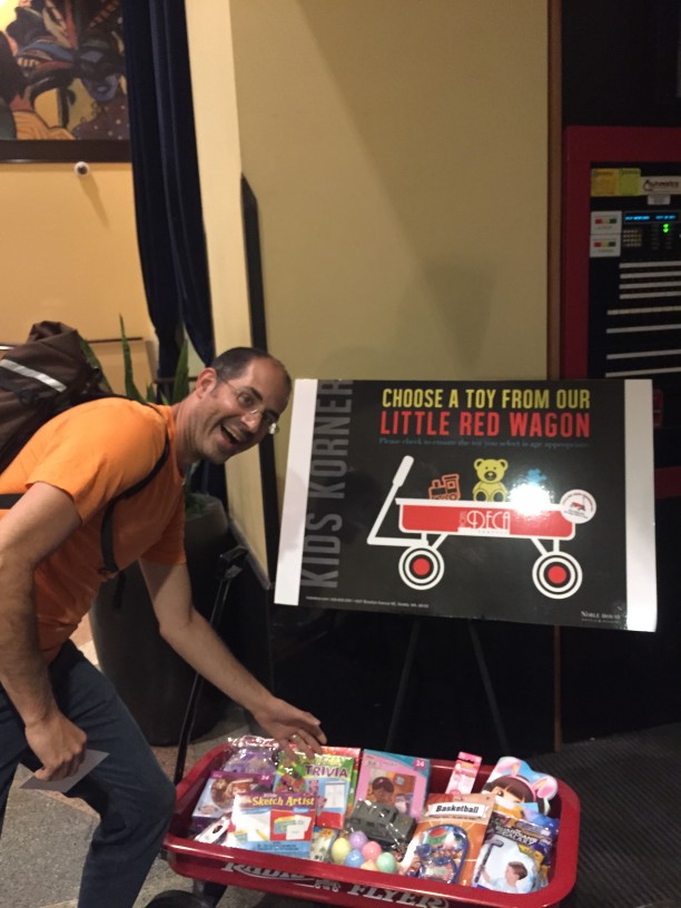 A person leaning a over a red wagon, full of toys, with a sign behind that says, Choose a toy from our little red wagon