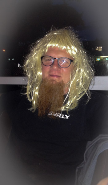 Headshot of a person with a long beard, wearing glasses and a blonde wig, in the dark