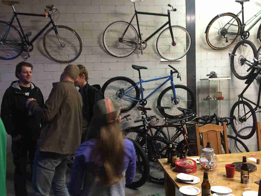 People standing in a room, next to a table, with Surly bikes mounted up on a cinder block wall behind them