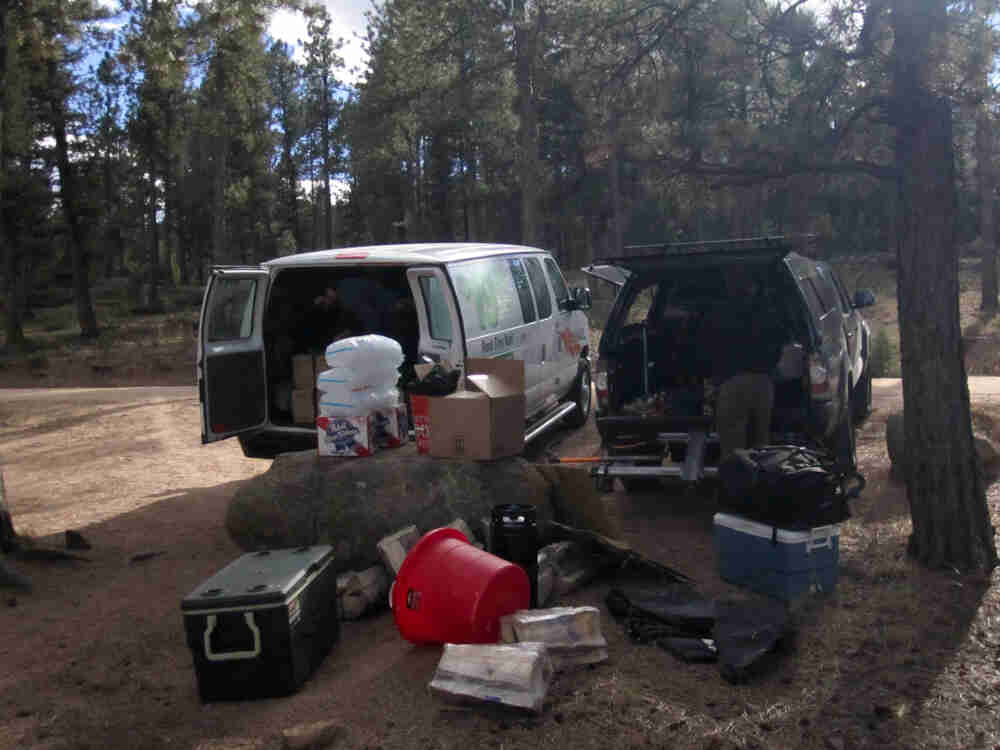 A pile of camping gear, in front of the rear of a van, and a truck with topper, with a forest in the background