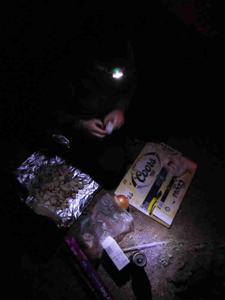 Downward view of a person wearing a headlamp, preparing food on the ground, at night