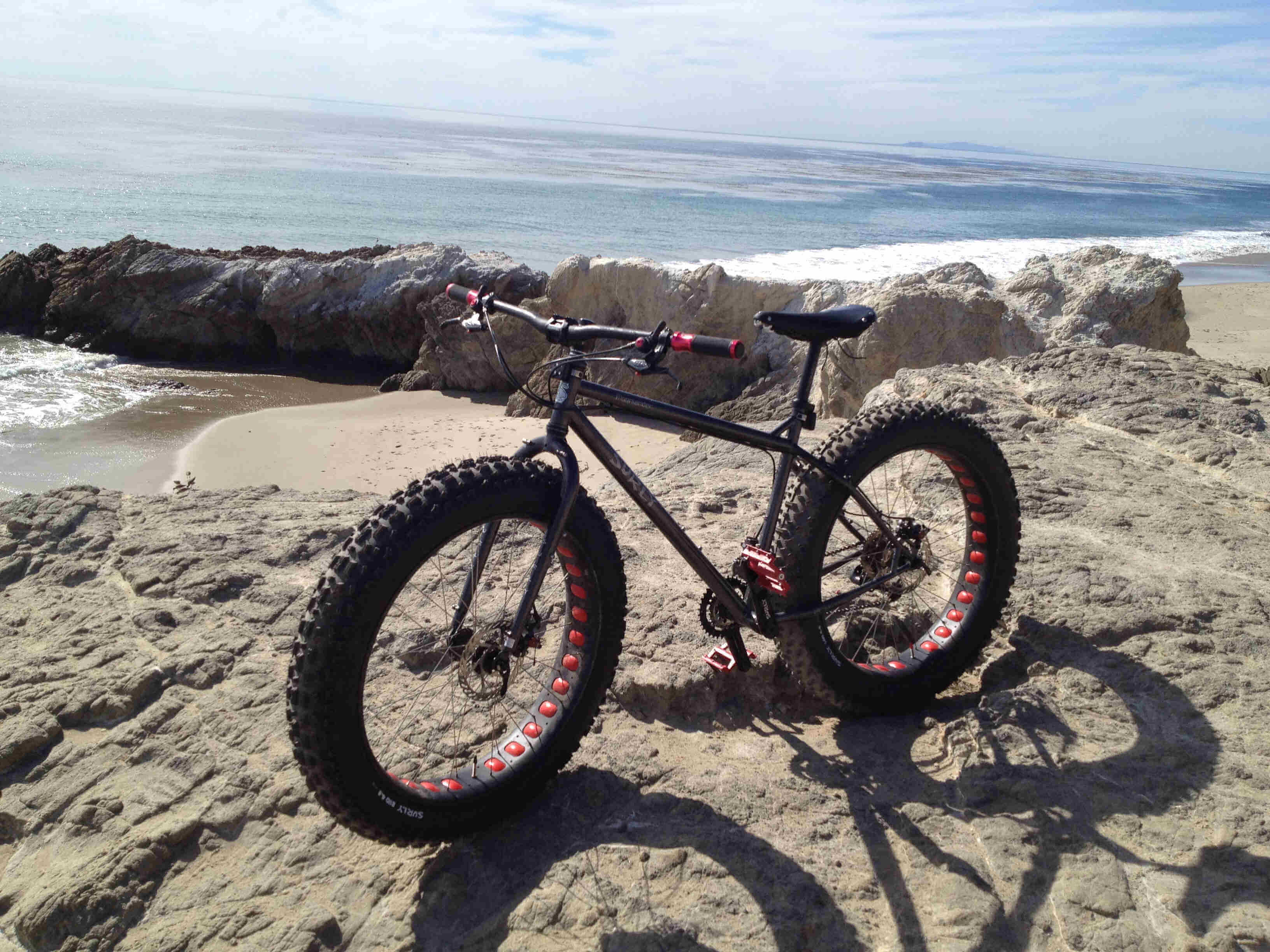 Left side view of a black Surly Moonlander fat bike, parked on a rock ledge, with the ocean behind