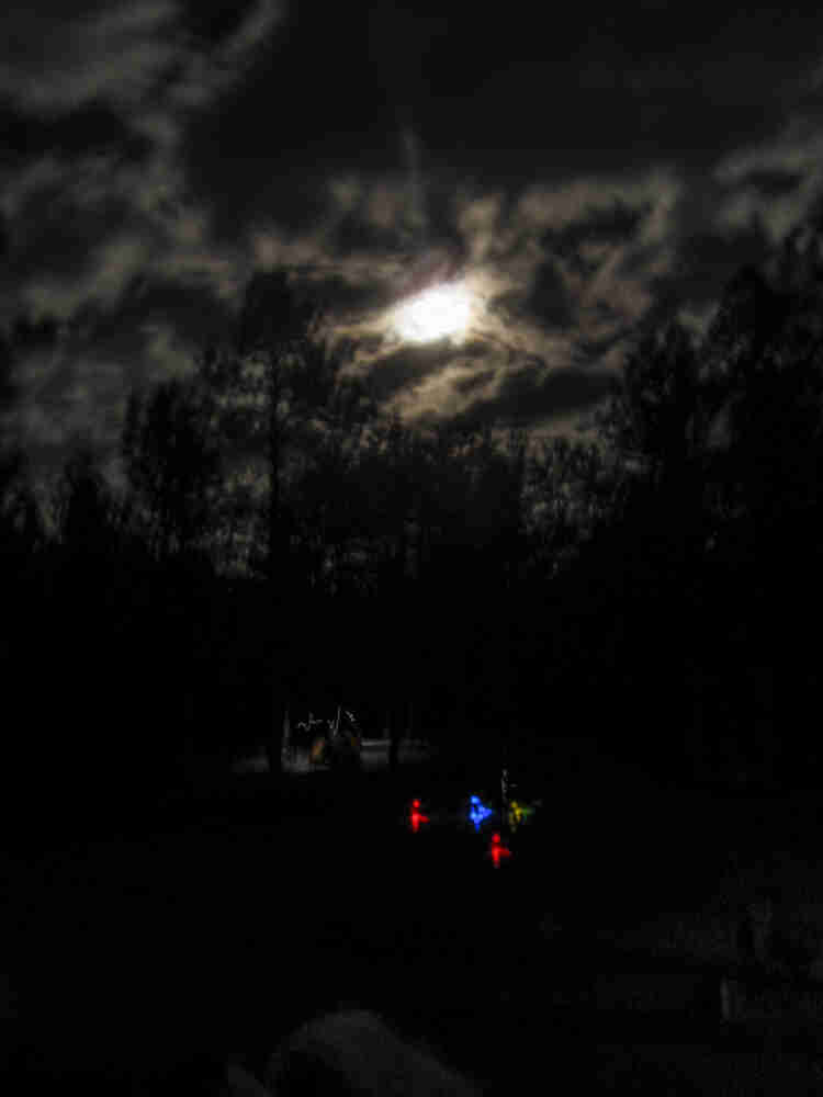 Colored lights lit up at night, with the moon shining through clouds above, with a campsite in the background