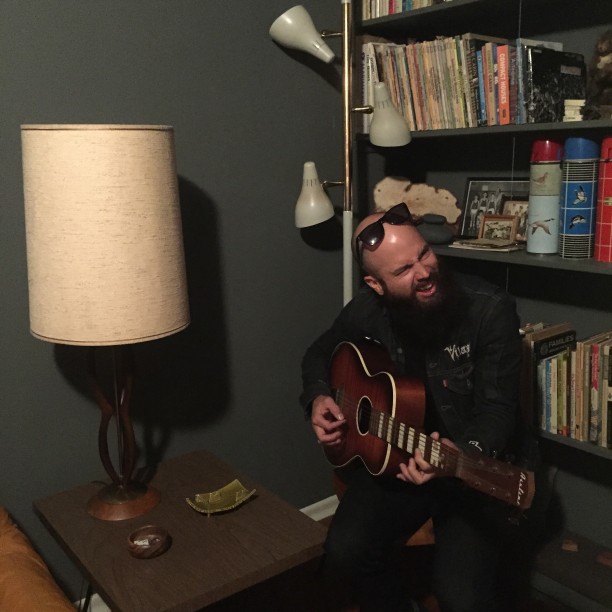 A person sitting in a chair, playing a guitar, next to a table and lamp, in front of a bookshelf