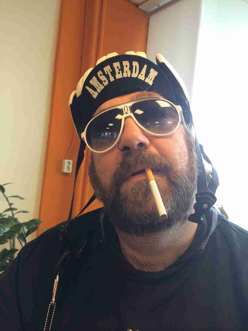Headshot of a person with a beard and a cigarette hanging from their mouth, wearing a bike helmet and sunglasses