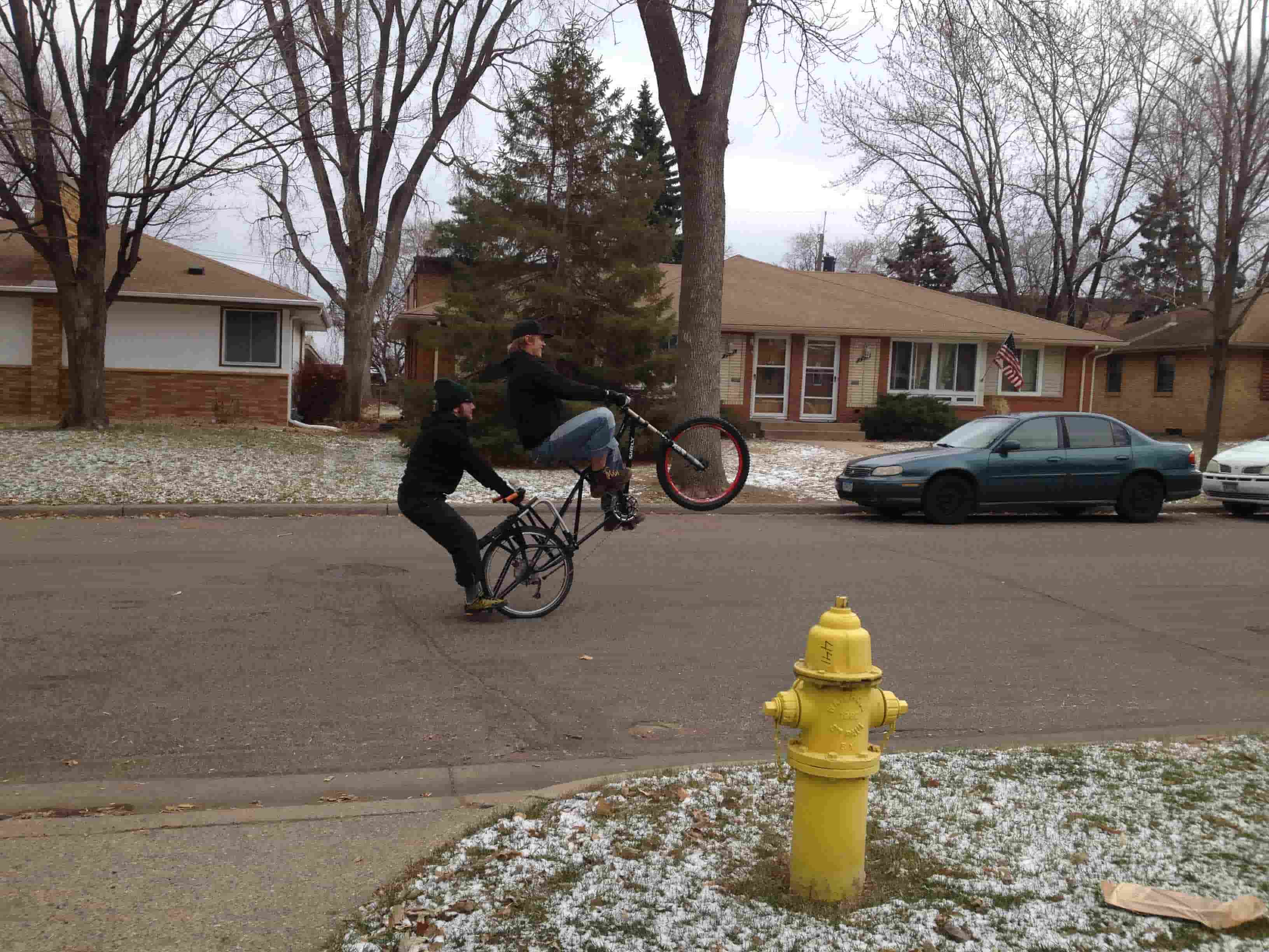 Right side view of a cyclist riding a wheelie on a black Surly Big Dummy bike with a person on back, on a paved street