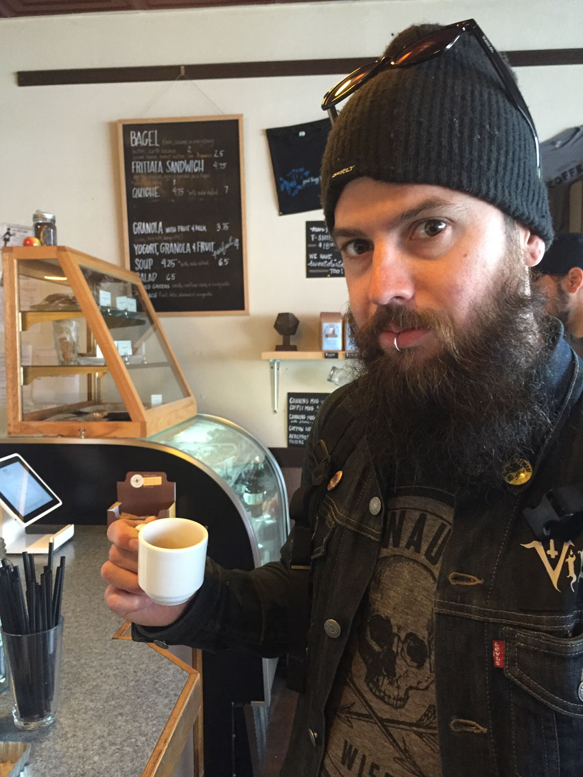Front view of a person posing with a cup of espresso, at the front counter of a cafe 