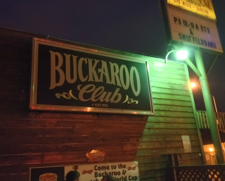 Wood sided wall of a building with a Buckaroo Club sign on it, at night with a light shining above