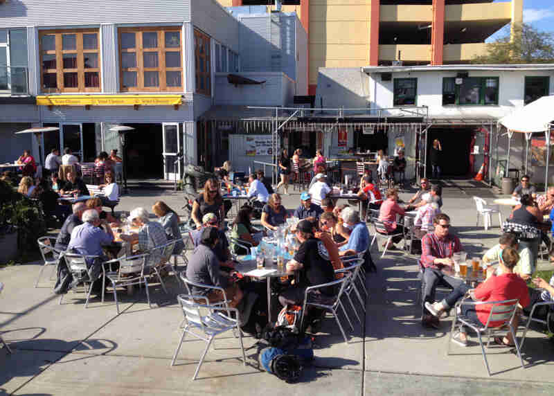 Several groups of people sitting at tables, on an outdoor patio area, in front of buildings with restaurants