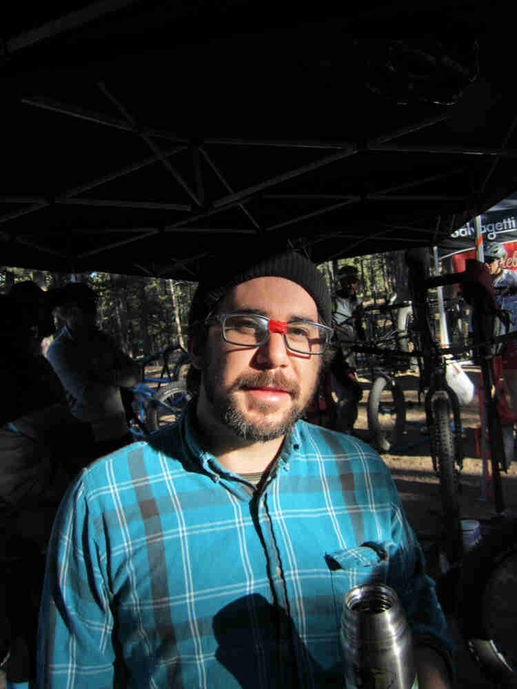 Front view of a person wearing glasses with red tape on the bridge, standing under a canopy with bikes and people