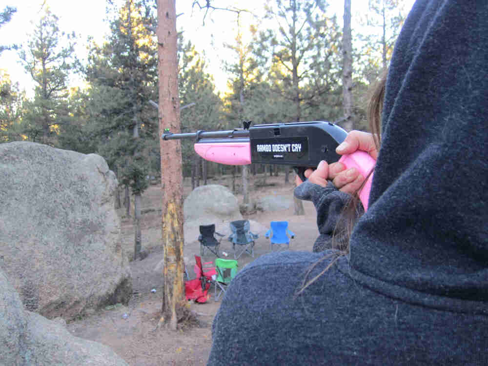 Rear view of a person wearing a hoodie, holding a rifle with a pink stock and butt, pointing towards the forest