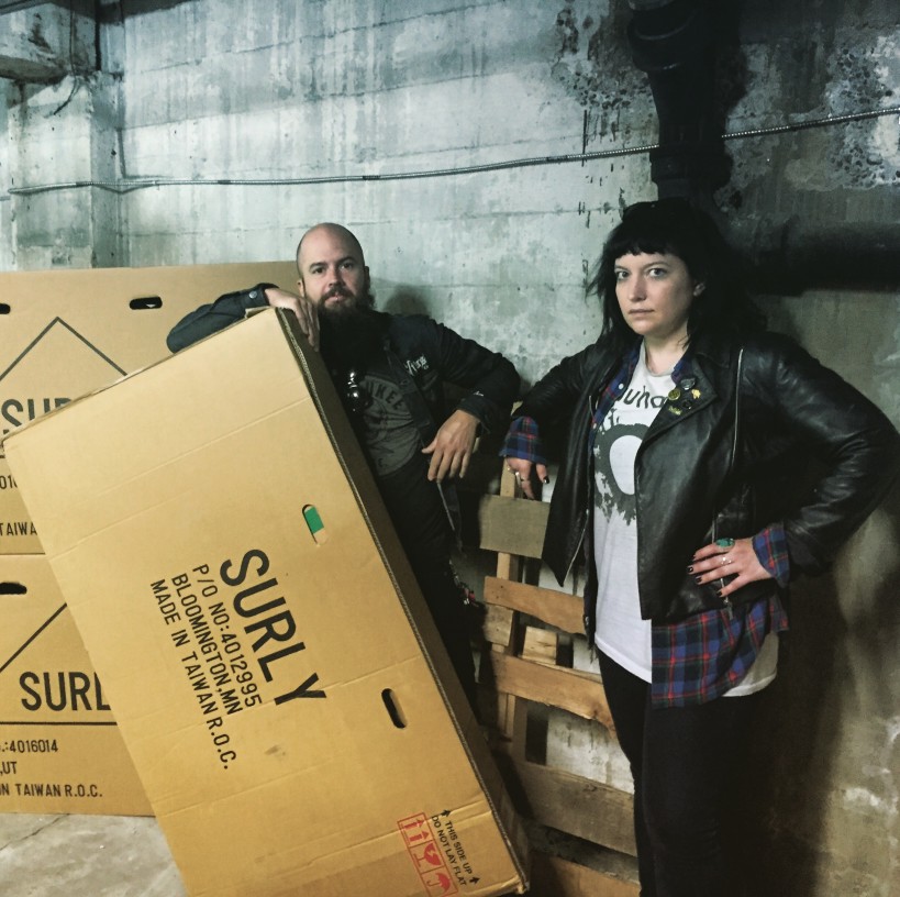 Front view of 2 people standing in a warehouse with cardboard Surly boxes next to them
