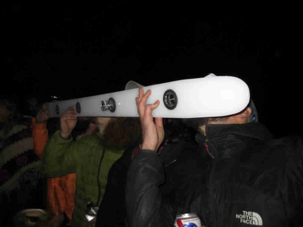 Nighttime with people taking shots from a snow ski, with holes in it for holding shot glasses