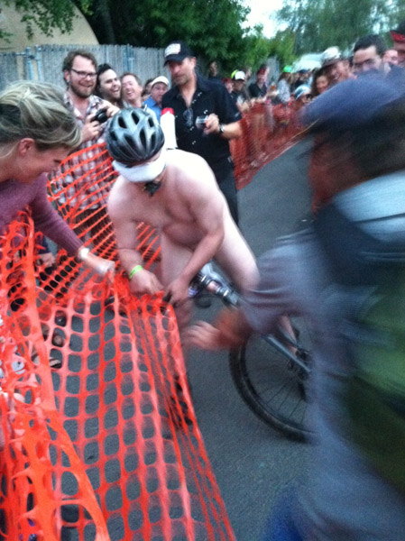 Front view of a cyclist wearing no clothes, falling into an orange snow fence, on a street with spectators watching