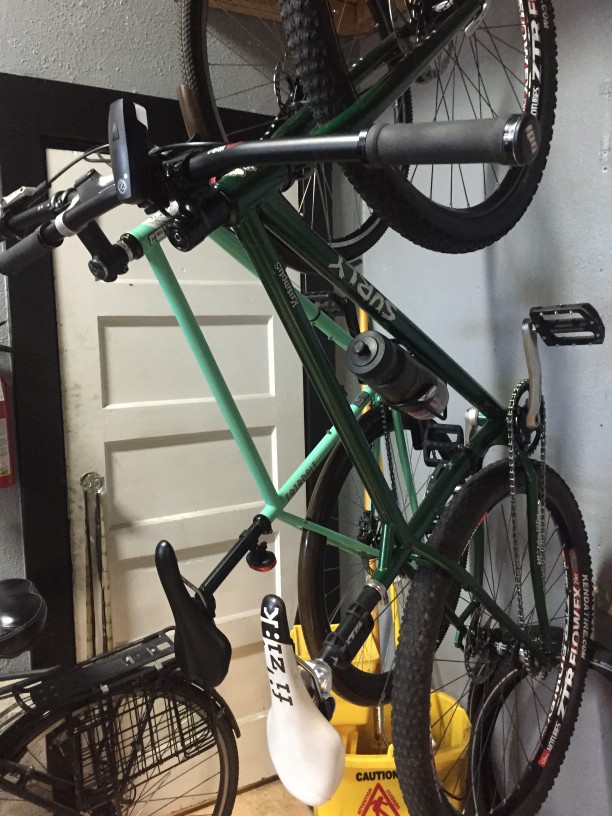 Right side view of a green Surly Krampus bike, hanging on a rack from the front wheel , in front a the door in a room
