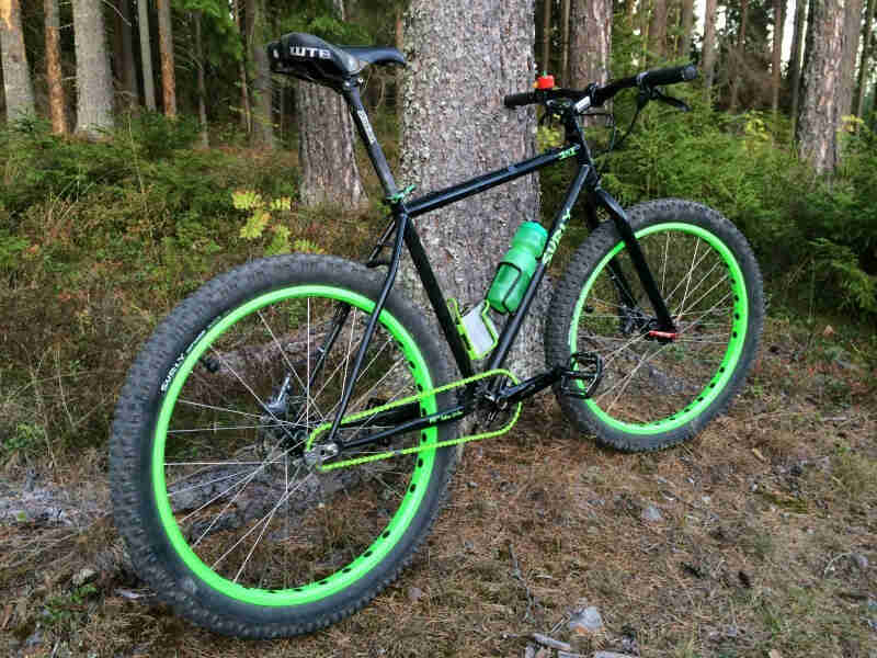 Right side view of a black Surly 1x1 bike with green rims, parked against a tree in a pine forest