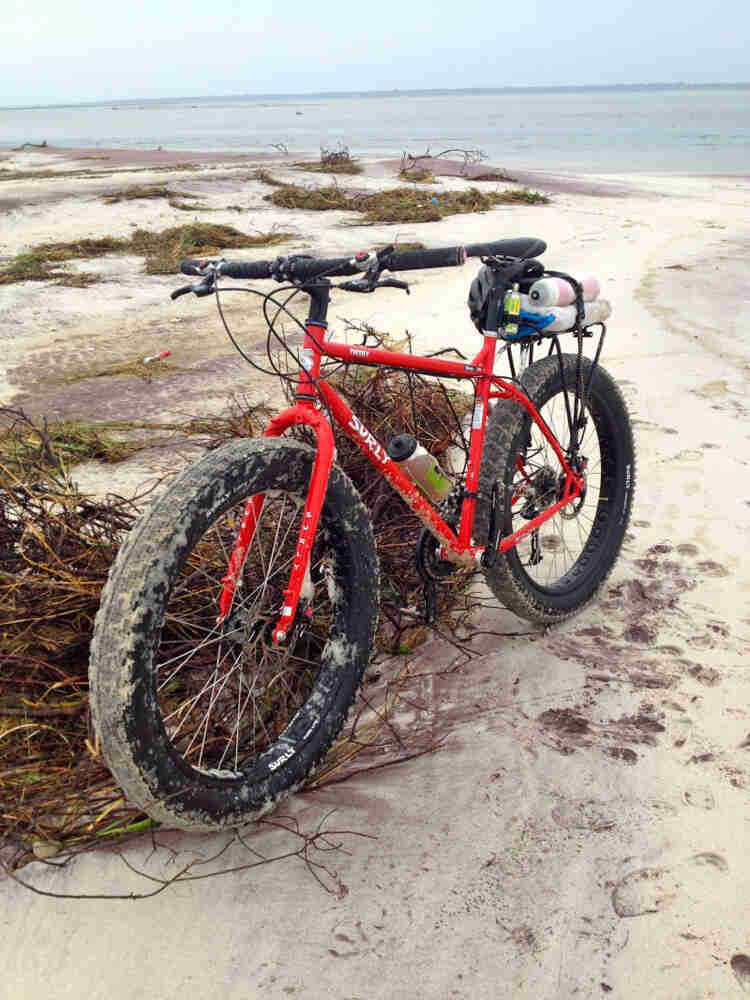 Front left view of a red Surly fat bike, on a sandy seashore with seaweed scattered around