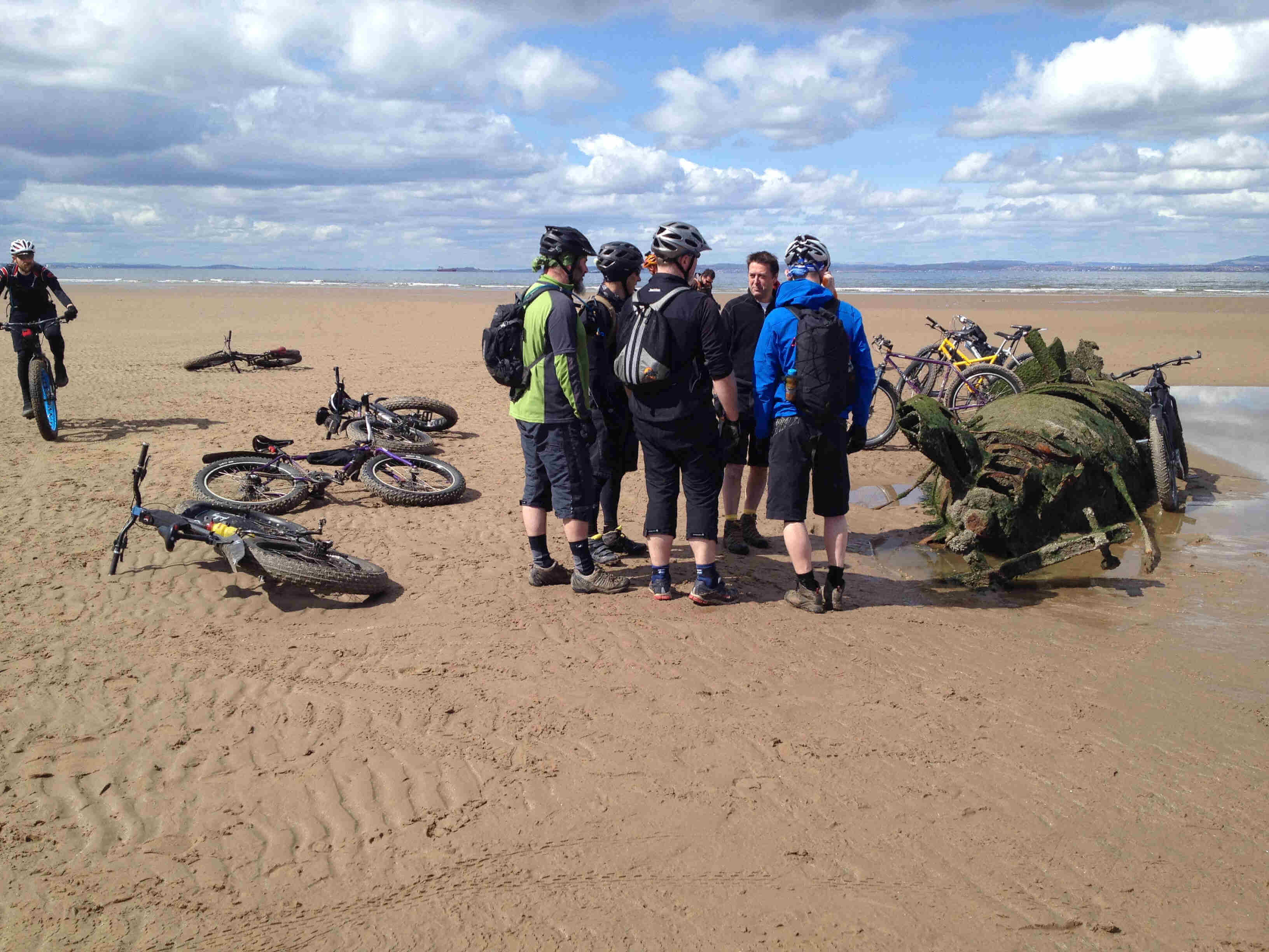 A group of cyclists standing in a circle with their Surly bikes laying around them, on a sandy area next to the ocean