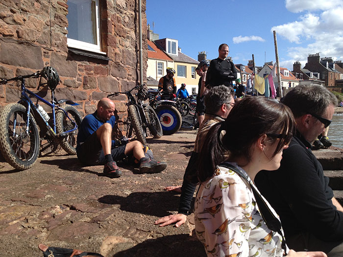 Right side view of people sitting on a stone walkway, with a blue Surly fat bike leaning on a stone building behind them