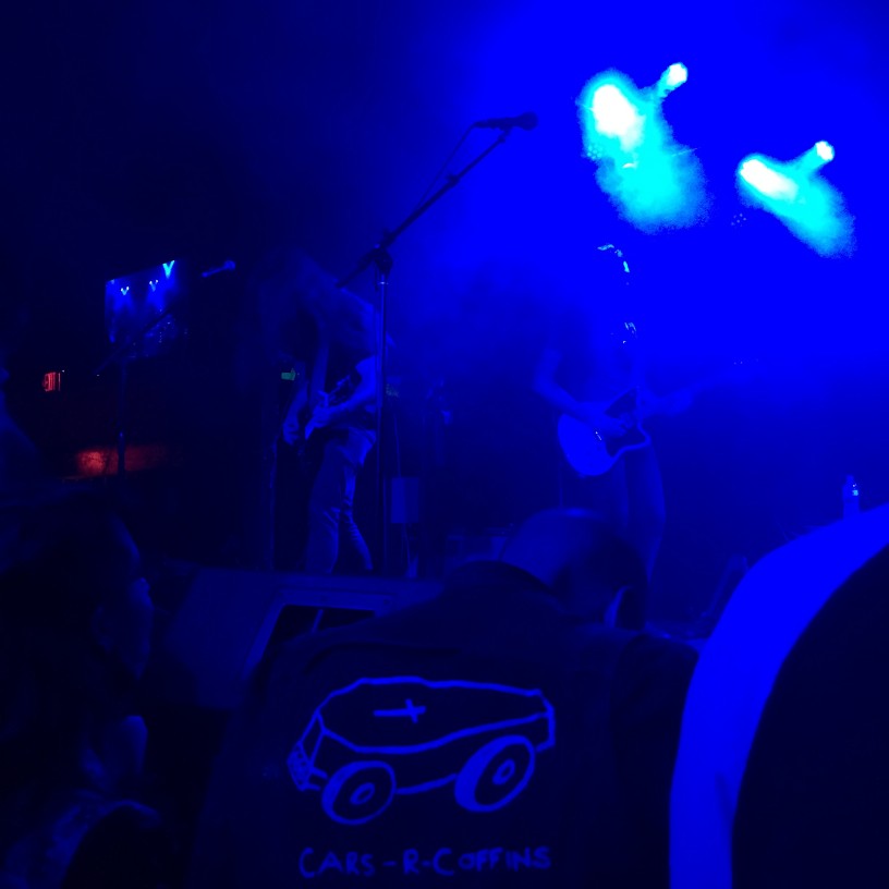 Rear view of people watching a band, up on a stage, in a dark room, with blue lights above