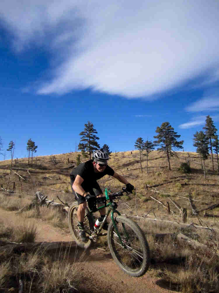 Front, right side view of a cyclist, riding a green Surly bike on a dirt trail, with a grassy hill in the background