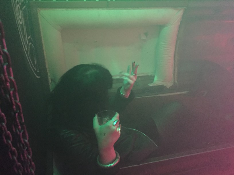 Downward view of person with long black hair, sitting in an open coffin with a beer in their hand