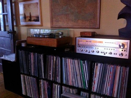 A 2 level cube shelf with a record player and stereo tuner on top, and albums underneath, against the wall inside a room