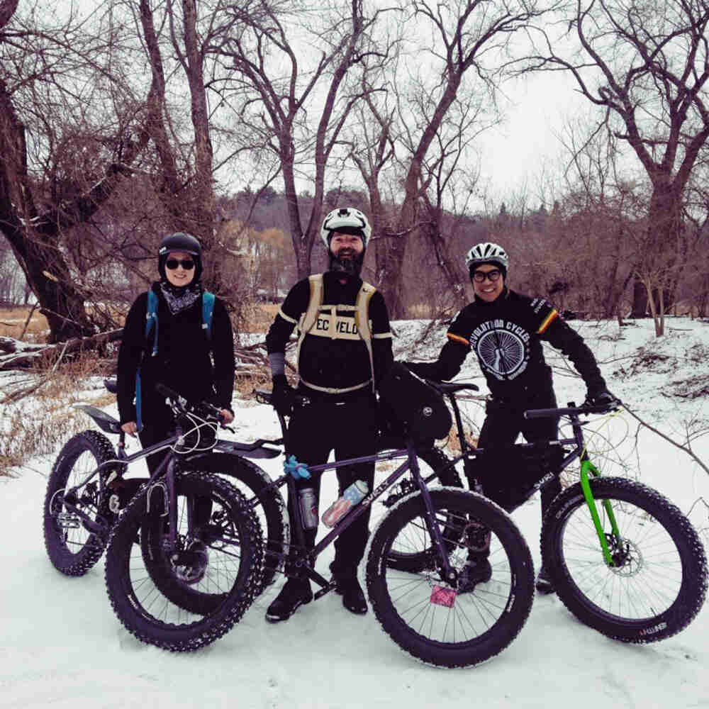 Front view of 3 cyclists standing with their Surly fat bikes on snow, with the woods in the background