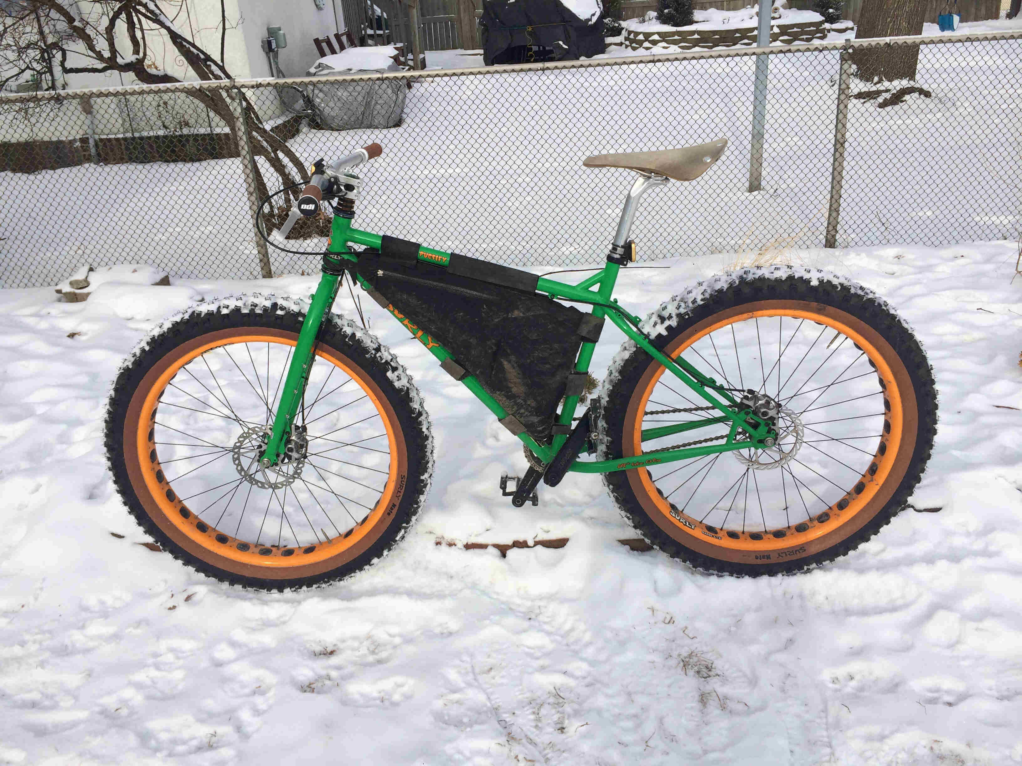 Left side view of a green Surly Pugsley fat bike with offset forks, parked in snow with a chain link fence in background