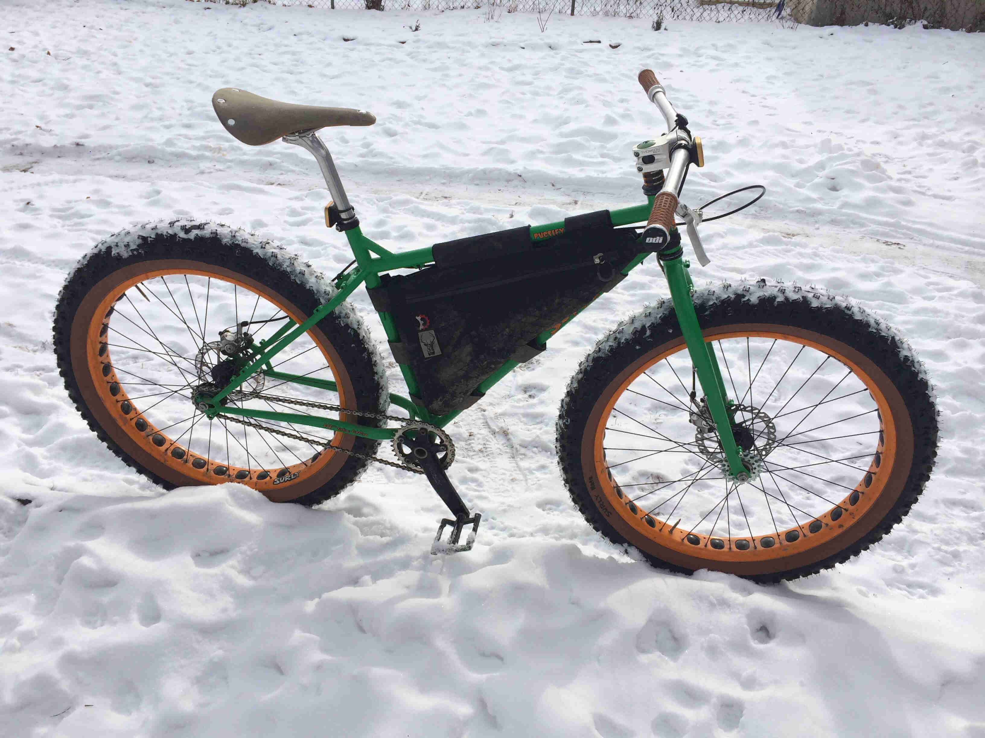 Right side view of a green Surly Pugsley fat bike with frame pack, parked along a snowbank on the side of a snowy trail